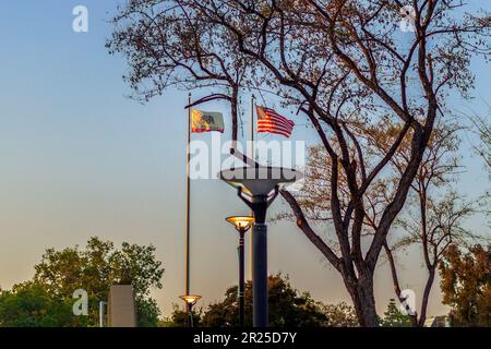 Ein inspirierendes Bild der goldenen Stunde von den US- und kalifornischen Flaggen, die hoch oben auf einem Flaggenmast am Crafton Hills College im Wind wehen. Stockfoto
