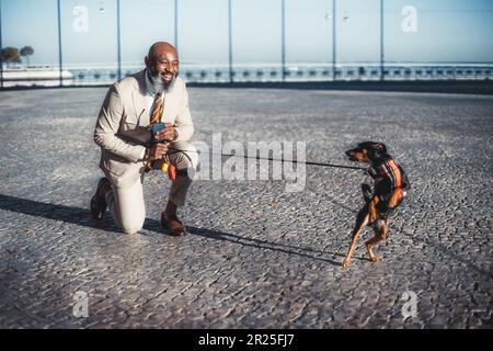 Selektive Fokussierung auf einen Mann mit kahlem Bart und einem Hund, hat ein Knie auf Lissabons Kopfsteinpflaster und lächelt den Hund an, der sich komisch benimmt; halt Stockfoto