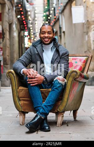 Ein afroamerikanischer Mann, der in die Kamera schaut und lächelt, während er draußen auf einem Stuhl sitzt. Stockfoto