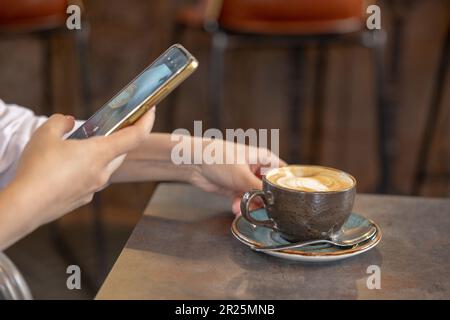 Ein junges Mädchen, das während der Aufnahme Fotos von Latte Art-Kaffee auf dem Display der mobilen Kamera macht. Hände einer Frau, die Kaffee fotografiert Stockfoto