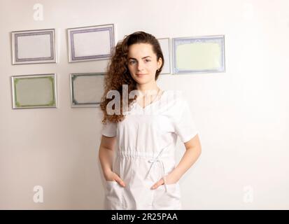Porträt Lächelnde Junge Weibliche Rehabilitationsspezialistin, Physiotherapeutin In Weißer Medizinischer Kleidung, Zertifikate In Rahmen An Der Wand Im Hintergrund Stockfoto