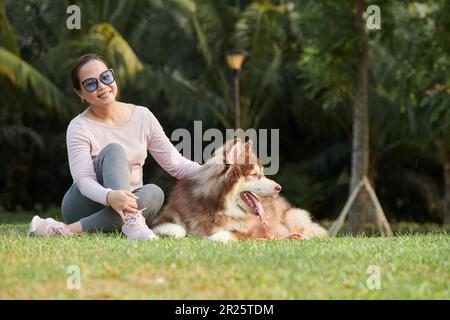 Eine glückliche Frau, die im Park auf dem Boden sitzt und ihren samoyierten Hund streichelt Stockfoto