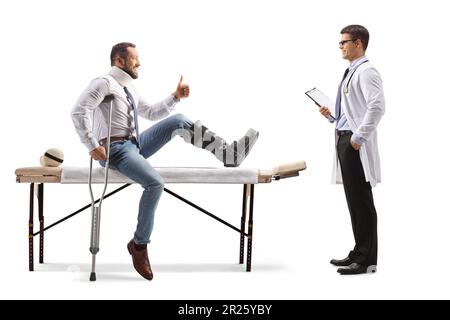 Verletzter Mann mit orthopädischem Stiefel und Halskragen, der auf einem Therapietisch sitzt und einem Arzt, isoliert auf weißem Hintergrund, Daumen hoch zeigt Stockfoto