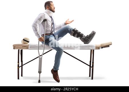 Verletzter Mann mit orthopädischem Stiefel und Halskragen, der auf einem Therapietisch sitzt und Gesten mit isolierter Hand auf weißem Hintergrund macht Stockfoto