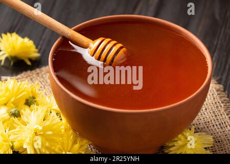 Honig in einer Tonschüssel, Dipper und gelbe Blumen auf Textil- und dunklem Holzhintergrund. Honigstäbchen liegen in einer Keramikschüssel mit Honignähe Stockfoto