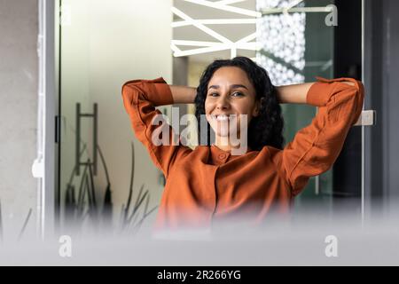 Zufrieden mit sich selbst und der geleisteten Arbeit sitzt eine junge lateinamerikanische Geschäftsfrau im Büro auf einem Stuhl und ruht sich mit den Händen hinter dem Kopf aus und lächelt. Stockfoto