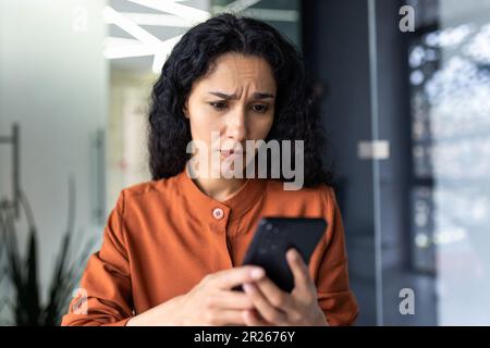 Verärgerte junge arabische Geschäftsfrau, die im Büro sitzt und sich Sorgen um das Telefon macht. Schlechte Nachrichten per Nachricht empfangen, Konto wird überprüft, Kommunikationsprobleme. Stockfoto