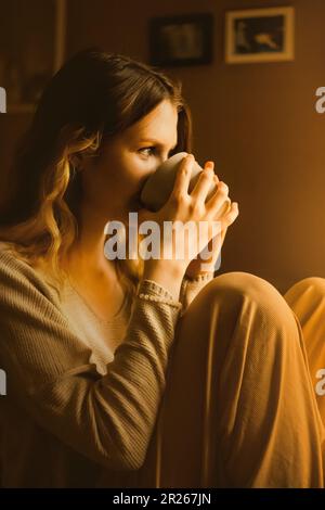 Eine zufriedene Dame, gekleidet in weicher beiger Loungewear, die zu Tagesbeginn eine Tasse Kaffee in der Küche isst. Sie führt eine ruhige Existenz, findet Vergnügen Stockfoto