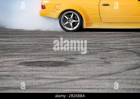 Nahaufnahme der Seitenansicht eines gelben Sportwagens, der auf einer grau geschwindigkeitsbetonten Asphaltstrecke driftet, wobei weißer Rauch aus dem Hinterrad austritt und Platz zum Kopieren vorhanden ist Stockfoto