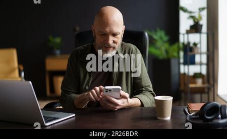 Glücklicher und charismatischer Senior-Mann, der sich in Textnachrichten am Telefon vertieft hat, während er mit dem Laptop eine Pause von der Arbeit machte. Seniorenmann, voller Vitalität, der den Komfort seines eigenen Hauses genießt. . Hochwertiges Foto Stockfoto