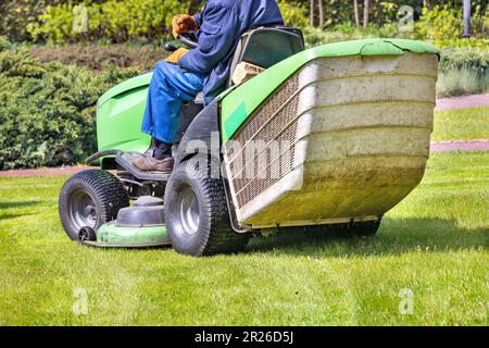 Ein Arbeiter in blauem Overall fährt einen Rasenmäher und mäht das grüne Gras des Rasens. Auf dem unscharfen Hintergrund eines Frühlingsgartens. Speicherplatz kopieren. Stockfoto