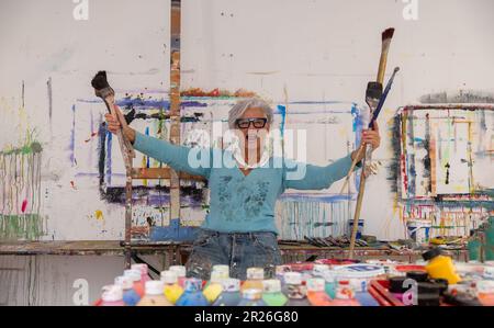Ältere grauhaarige, reife, fröhliche, lachende Künstlerin mit Brille und großen Pinseln in ihren Händen, im Studio, Kopierraum Stockfoto