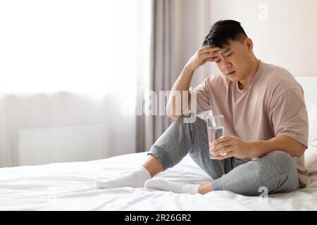 Asiatischer Mann mit Schmerzen, der auf dem Bett sitzt und Glaswasser hält Stockfoto