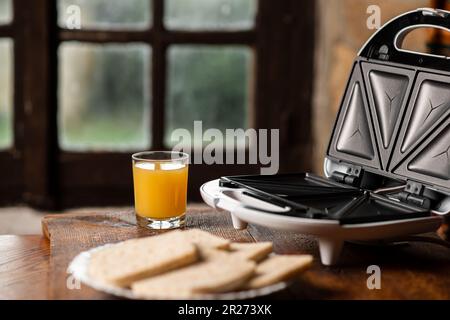 Sandwichmaker und ein Glas Saft auf dem Hintergrund eines altmodischen Fensters. Ich bereite ein schnelles Frühstück und einen Snack vor. Stockfoto