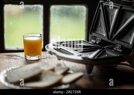 Sandwichmaker und ein Glas Saft auf dem Hintergrund eines altmodischen Fensters. Ich bereite ein schnelles Frühstück und einen Snack vor. Stockfoto