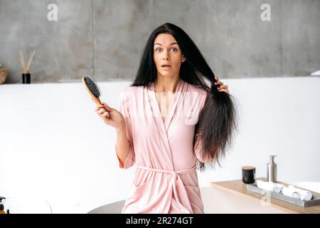 Wunderschöne überraschte weiße Frau in einem Bademantel, mit dicken, langen schwarzen Haaren, sitzt im Badezimmer, hält einen Kamm in der Hand und schaut nach dem Haarbürsten erstaunlich auf die Kamera Stockfoto