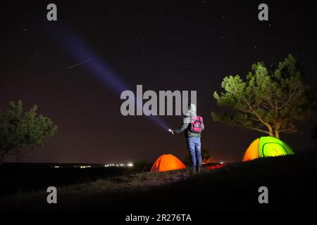 Ein Mann mit heller Taschenlampe in der Nähe von Campingzelten im Freien bei Nacht Stockfoto