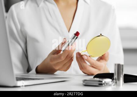 Junge Frau mit Kosmetikspiegel und Lippenstift am weißen Tisch in geschlossenen Räumen, Nahaufnahme Stockfoto