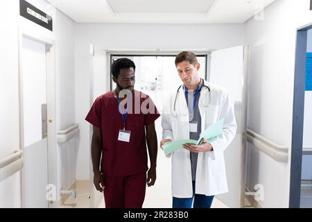 Zwei verschiedene männliche Ärzte schauen auf Notizen und gehen im Krankenhausflur Stockfoto