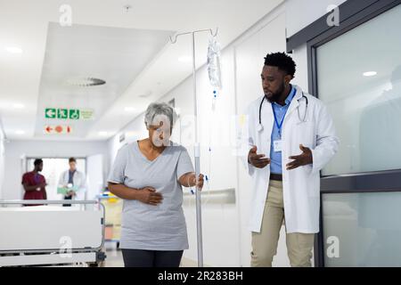 Ein afroamerikanischer Arzt, der mit einer vielschichtigen älteren Patientin spricht, die einen Tropfstand im Korridor hält Stockfoto