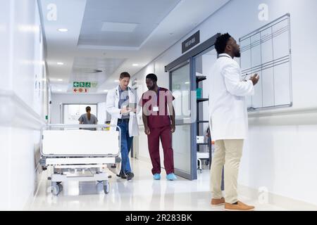 Zwei verschiedene männliche Ärzte, die Tablet benutzen und sprechen in einem belebten Krankenhausflur Stockfoto