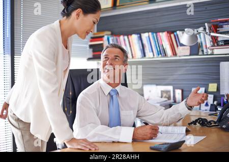 Er hätte sich keinen besseren Assistenten wünschen können. Ein erwachsener Geschäftsmann, der mit einer Mitarbeiterin spricht, während er an seinem Schreibtisch sitzt. Stockfoto
