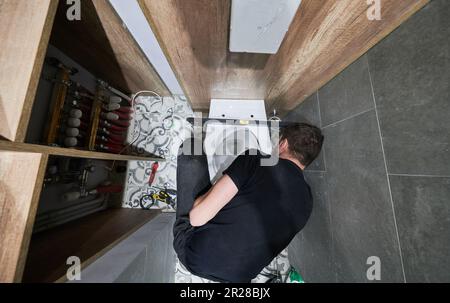 Draufsicht eines Mannes, der eine Toilettenschüssel in einem Apartment installiert, das gerade renoviert wird. Männlicher Klempner, der sich mit Bauwerkzeugen auf dem Boden niederhockt, während er zu Hause an der Toiletteninstallation arbeitet. Stockfoto