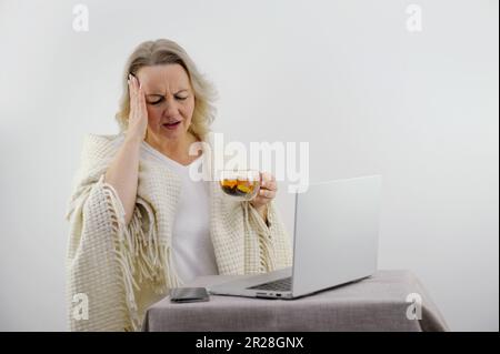 Das kranke brünette Mädchen sitzt auf dem Sofa und arbeitet an einem Laptop. Die Frau hat Rotz, Fieber und Kopfschmerzen. Junge Frau wird zu Hause behandelt, hat sich krankgeschrieben. Winterkälte und Grippe Stockfoto