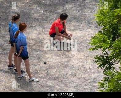 Drei junge Mädchen mit Petanque-Bällen, die darauf warten, auf einem Sandboden-Petanque-Platz zu spielen. Ein Mädchen hockt und die anderen zwei stehen auf einem sonnigen Vater Stockfoto