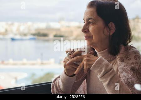 Eine reife Frau mittleren Alters, die eine Tasse Tee oder Kaffee hält, und während des Tages pensiv aus einem großen, hellen Fenster schaut Stockfoto