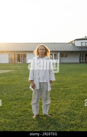 Ein Yoga-Mann in Baumwollkleidung, der auf einem grasbedeckten Rasen neben dem Cottage im Hintergrund steht Stockfoto