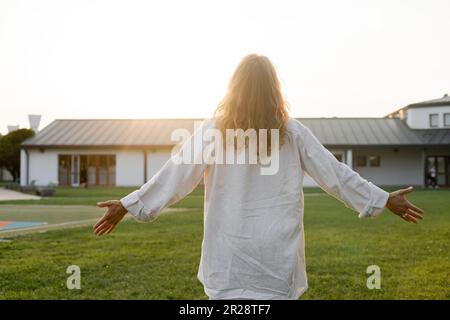 Rückansicht eines langhaarigen Mannes im weißen Leinenhemd, der mit ausgestreckten Händen in der Nähe einer Hütte im Freien meditiert Stockfoto