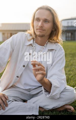 Ein Mann mit langen Haaren in Leinenbekleidung, der mala-Perlen berührt und während der Meditation im Freien wegschaut Stockfoto