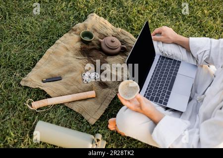 Teilansicht eines Mannes mit Laptop, der Tonbecher in der Nähe von Teekanne und mala Perlen auf Leinenteppich auf grünem Rasen hielt Stockfoto