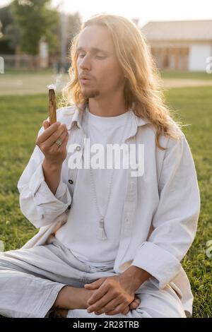 Ein langhaariger Mann in weißer Leinenbekleidung bläst während eines esoterischen Rituals im Freien auf den aromatischen palo santo Stab Stockfoto