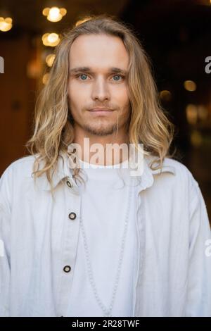 Porträt eines blonden Yoga-Mannes im weißen Hemd, der in die Kamera schaut, mit verschwommener Stadt im Hintergrund Stockfoto