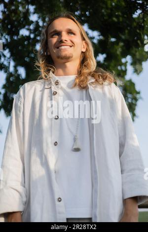 Blick aus dem niedrigen Winkel auf einen sorgenfreien Yoga-Mann mit langen Haaren in Leinenbekleidung, der draußen lächelt Stockfoto