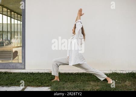 Seitlicher Blick auf einen barfuß Mann in weißer Baumwollkleidung, der in der Krieger-Pose meditiert, mit erhobenen Gebetshandbeinen in der Nähe eines modernen Cottage Stockfoto