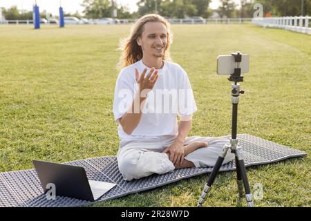 Fröhlicher Yoga-Coach, der in bequemer Pose neben Laptop und Mobiltelefon auf einem Stativ auf dem grünen Rasen des Stadions sitzt Stockfoto