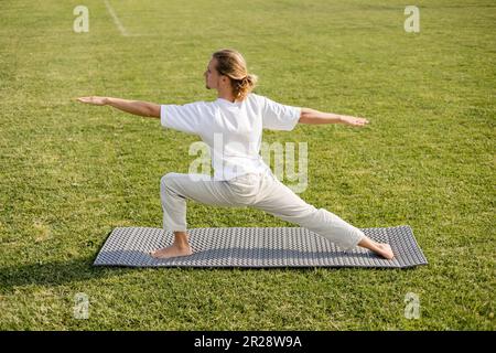 Seitenansicht eines jungen, langhaarigen Mannes im weißen T-Shirt, der in der Krieger-Pose meditiert, mit ausgestreckten Händen auf der Yogamatte im Freien Stockfoto