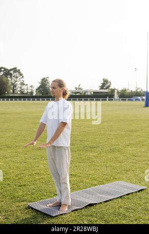 Ein langer Mann in Leinenhosen und ein weißes T-Shirt meditieren mit geschlossenen Augen, während er auf einer Yoga-Matte auf grünem Rasen steht Stockfoto