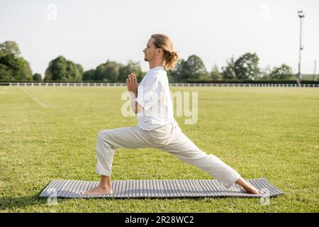 Seitenansicht eines Barfußmanns, der Yoga in Krieger-Pose übt, mit anjali-Mudra-Geste auf grünem Gras im Freiluftstadion Stockfoto