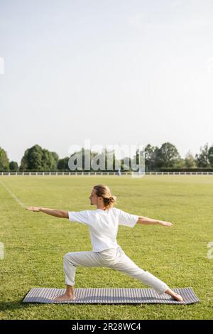 Seitenansicht eines langhaarigen und barfuß-Mannes, der Yoga in der Krieger-Pose mit ausgestreckten Händen auf dem grasbedeckten Feld übt Stockfoto