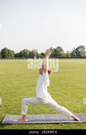 Seitenansicht eines Mannes in Leinenhosen und einem weißen T-Shirt, der mit erhobenen Händen auf Yoga-Matte auf grünem Rasen eine Krieger-Pose übt Stockfoto