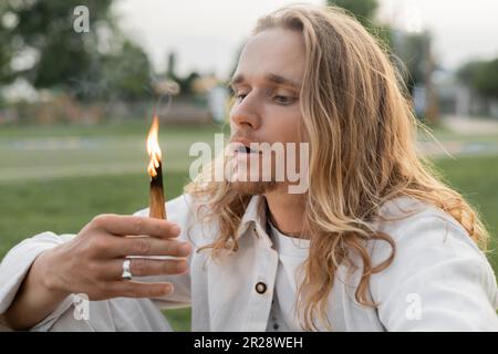 Ein langhaariger Mann in weißen Klamotten bläst palo-santo-Stäbchen in die Luft Stockfoto