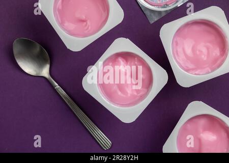 Joghurt mit Fruchtgeschmack in weißen Plastikbechern auf hellem lila Hintergrund mit Deckel aus silberner Folie – Überkopffoto des Joghurtbechers mit selektivem Hintergrund Stockfoto
