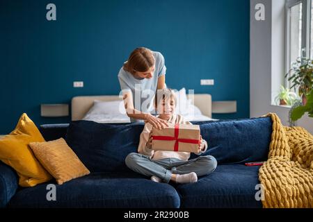 Mutter begrüßte den kleinen Sohn mit Geburtstag, schenkt Geschenkbox, sitzt auf dem Sofa im Wohnbereich Stockfoto