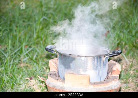 Kochen Sie Suppe in einem Edelstahltopf. Das Abendessen ist heiß auf dem Holzkohlegrill. Weißer Rauch von der Hitze. Gras verschwommener Hintergrund. Nahaufnahme und selektiv für Stockfoto