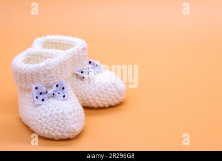 Gestrickte weiße Babyschühchen mit Schleifen in schwarzen Punkten auf einem zarten, orangefarbenen, isolierten Hintergrund. Hintergrund. Für Text. Foto in hoher Qualität Stockfoto