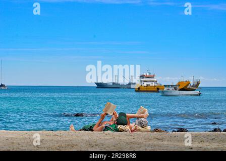 Ein Paar, das am Playa Mann Beach liest. Schiffe im Hintergrund. Die Insel San Cristobal. Galapagosinseln, Ecuador Stockfoto